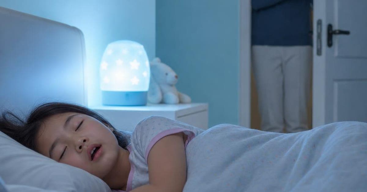 Young child asleep with mouth open under a soft nightlight while a parent watches from the doorway